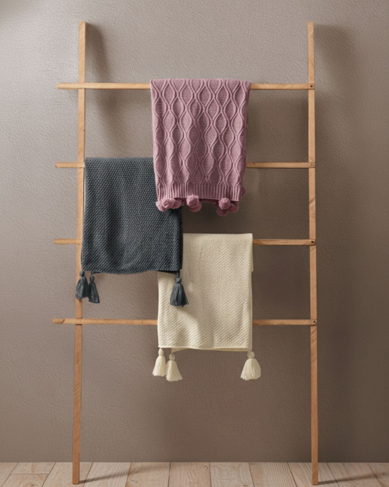 A wooden ladder leans against a wall, displaying three knitted blankets: a pink frazada ligera with diamonds and pompones, a gray one with airy knit and tassels, and the cream Frazada de Lujo Chai Beige by Intima Hogar, all on a wooden floor.
