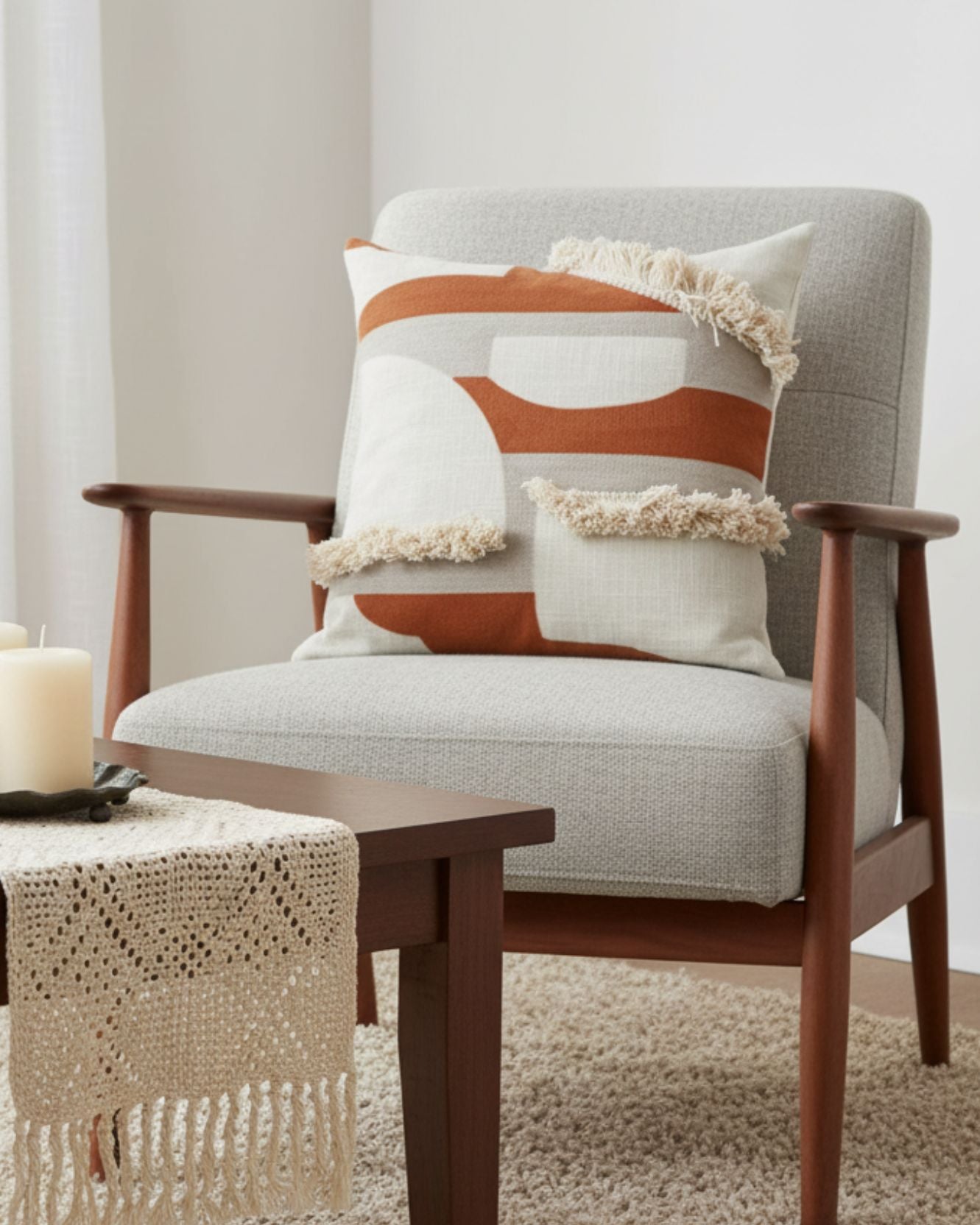 A modern armchair with light gray upholstery and wooden arms holds two geometric pillows, including the Funda de Cojín Agadir by Intima Hogar. In front, a wooden table with candles and a lace runner rests on a beige carpet.