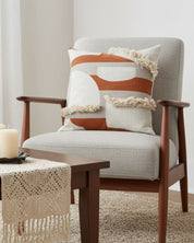 A modern armchair with light gray upholstery and wooden arms holds two geometric pillows, including the Funda de Cojín Agadir by Intima Hogar. In front, a wooden table with candles and a lace runner rests on a beige carpet.