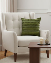 A modern beige armchair with the Intima Hogar Funda de Cojín Bogotá in green textured fabric sits beside a wooden coffee table with candles and a small vase in a cozy, softly lit living room.