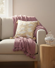 A beige sofa with a mauve textured throw, pom-poms, the Funda de Cojín Casablanca by Intima Hogar, a glass jar of candies on a wooden side table, and soft natural light create un espacio sereno in this cozy living room.
