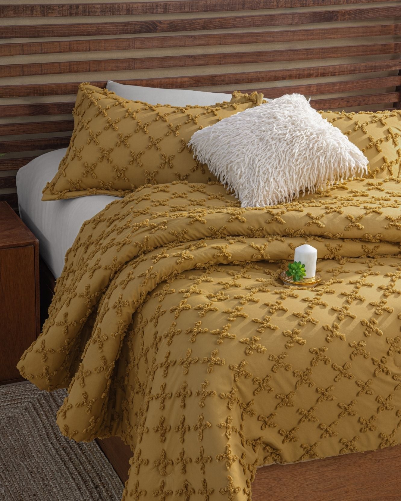 A bed dressed in mustard yellow bedding with the Funda de Cojín Kawasaki from Intima Hogar and a white shaggy pillow exudes relaxed luxury. A bedside table with a candle and succulent completes the look beneath a slatted wood headboard.