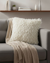 A fluffy white pillow with the Funda de Cojín Kawasaki by Intima Hogar rests on a light gray sofa draped with a beige knit blanket, while a wooden shelf with books and minimalist vases decorates the wall behind.