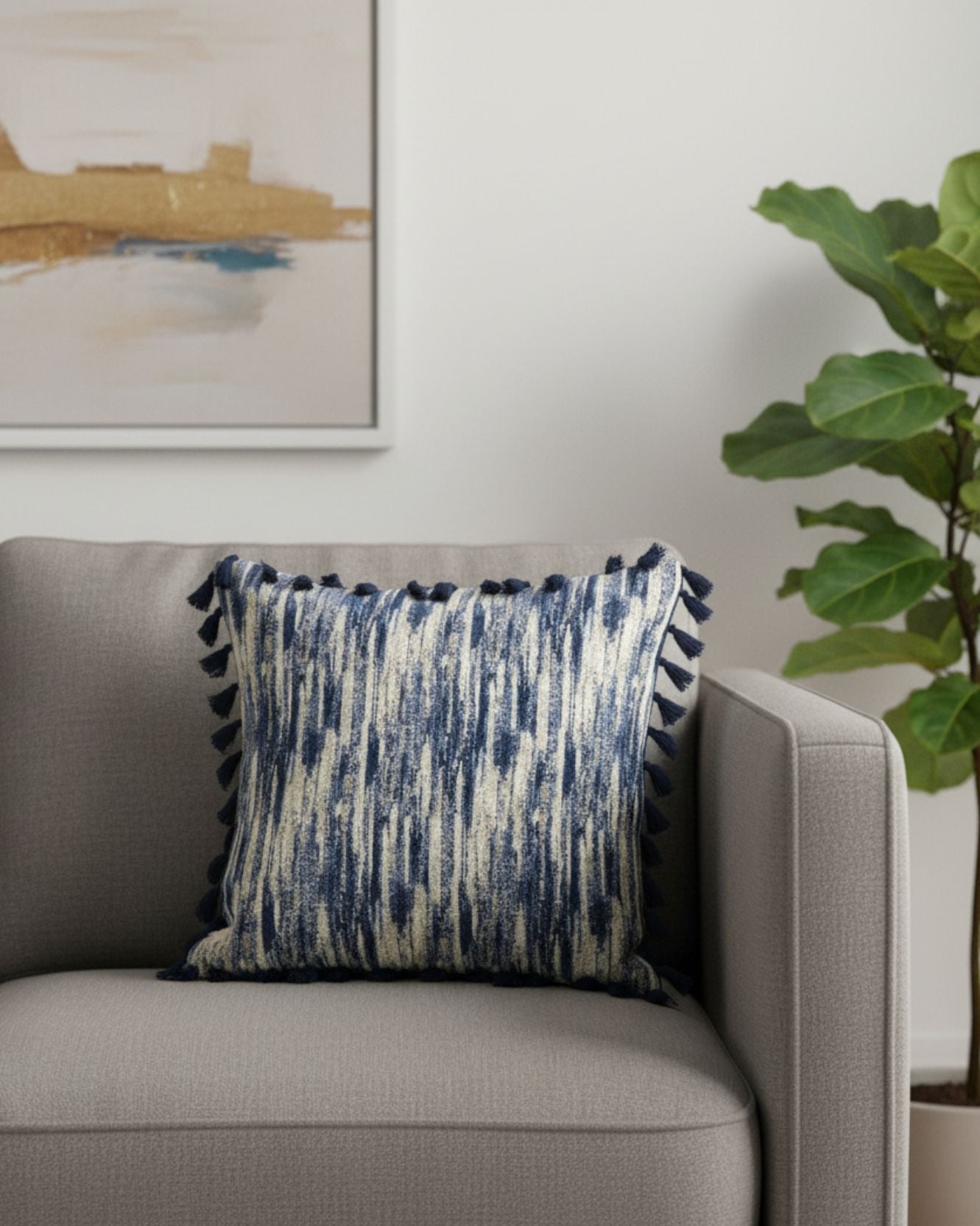 La funda de cojín Marrakech de Intima Hogar, con un diseño azul y crema y borlas, decora un sillón gris claro. Una planta verde y arte abstracto completan un ambiente acogedor y moderno con decoración elegante.