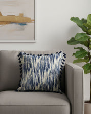 La funda de cojín Marrakech de Intima Hogar, con un diseño azul y crema y borlas, decora un sillón gris claro. Una planta verde y arte abstracto completan un ambiente acogedor y moderno con decoración elegante.