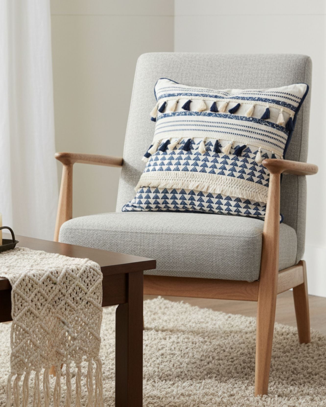 A modern light gray armchair with wooden accents features the Funda de Cojín Meknes by Intima Hogar. A brown table with a crochet runner and beige rug complete this cozy, softly lit room de inspiración hogar natural.