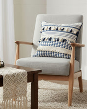 A modern light gray armchair with wooden accents features the Funda de Cojín Meknes by Intima Hogar. A brown table with a crochet runner and beige rug complete this cozy, softly lit room de inspiración hogar natural.