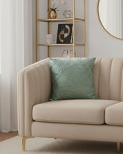 A modern beige sofa with the light green Funda de Cojín Osaka by Intima Hogar sits before a gold shelving unit and round mirror in a bright living room with white curtains, neutral decor, and unique interior design details.