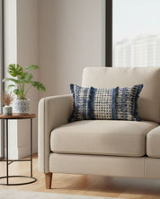 A beige jaspeado armchair with the Funda de Cojín Rabat by Intima Hogar in blue and white sits beside a round side table with a potted plant, vase, and dried flowers, adding a cozy touch to the bright room with large windows.