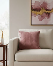 A light beige armchair with the Intima Hogar Funda de Cojín Sakai pillow sits by a wooden side table. Above, a pink and gold abstract painting hangs on a light wall.