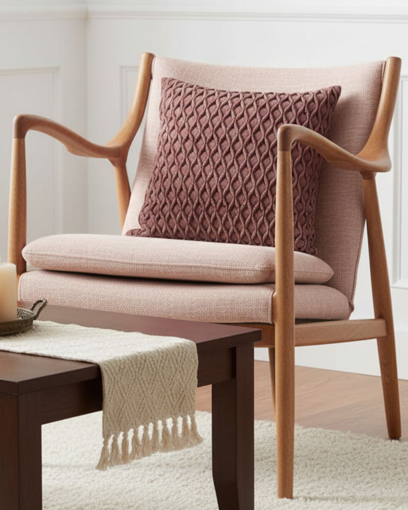 The Funda de Cojín Sendai by Intima Hogar in mauve adds textured style to a modern wooden armchair with pink upholstery, behind a dark wood coffee table with a cream runner and candles, set against white paneled walls and decor moderno.