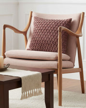The Funda de Cojín Sendai by Intima Hogar in mauve adds textured style to a modern wooden armchair with pink upholstery, behind a dark wood coffee table with a cream runner and candles, set against white paneled walls and decor moderno.