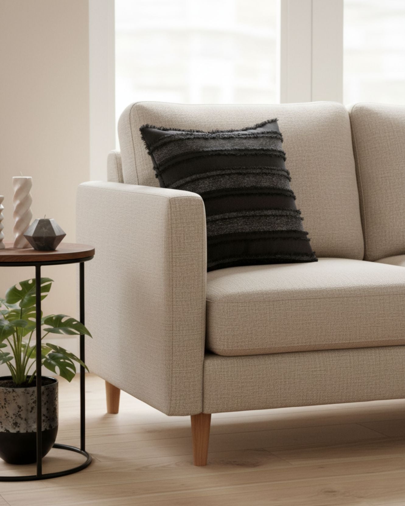 A modern beige sofa with the Funda de Cojín Seúl by Intima Hogar in black adds un toque de decoración atractivo, next to a round side table with a small plant and decor, in a bright room with large windows and light wood floors.