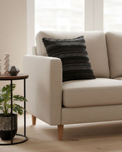 A modern beige sofa with the Funda de Cojín Seúl by Intima Hogar in black adds un toque de decoración atractivo, next to a round side table with a small plant and decor, in a bright room with large windows and light wood floors.