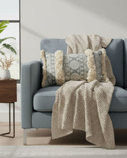A light blue sofa is styled with a textured beige throw blanket featuring franjas con flecos and two Intima Hogar Funda de Cojín Yadida pillows. Nearby, a wooden side table holds a vase of dried flowers and a leafy plant by the sunlit window.
