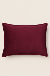 La Funda de Almohada Ensueño Vino de Intima Hogar, rectangular con textura aterciopelada y brillo suave en color burdeos, destaca sobre fondo beige. Su funda removible y borde sencillo añade elegancia a tu cama o habitación.