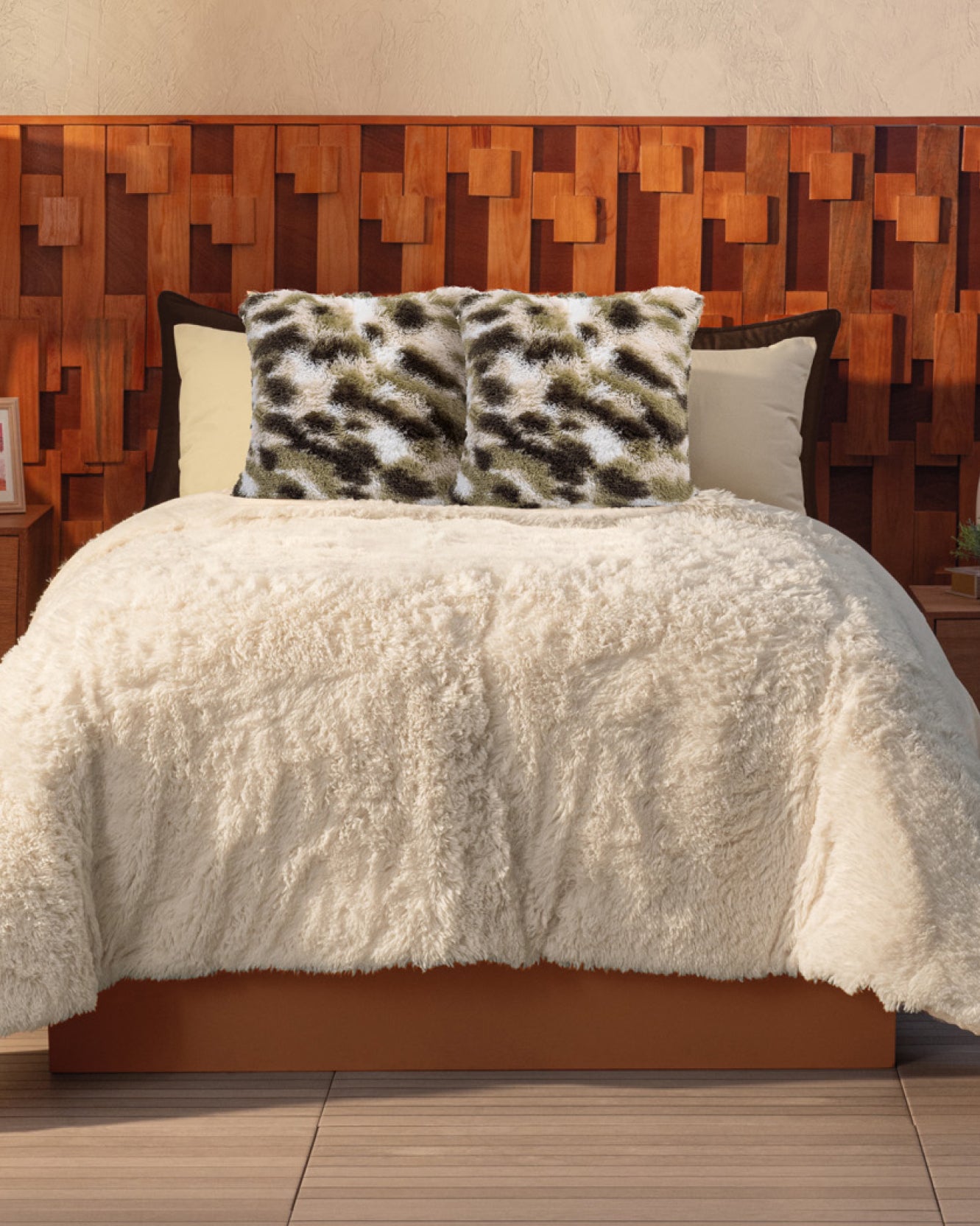 A bed with the Cobertor Platinum Extra Suave Iglú by Intima Hogar and two patterned fuzzy pillows sits against a textured wooden accent wall, while nightstands with framed photos complete this cozy iglú crema retreat.