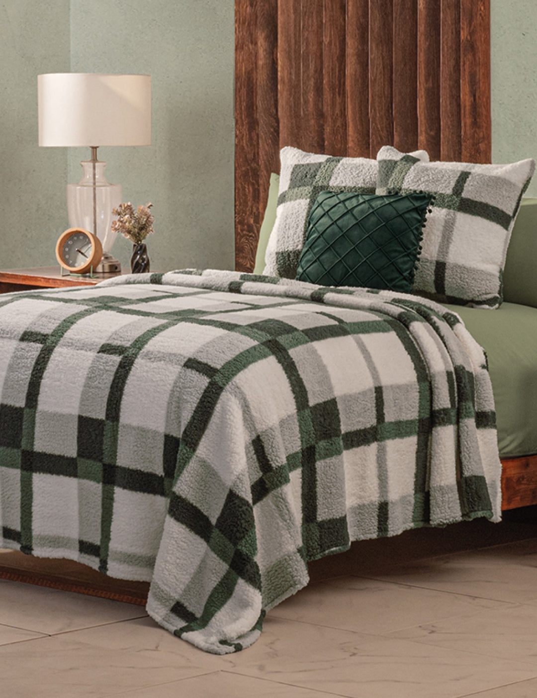 A neatly made bed features the Intima Hogar Juego de Cobertor Flannel Ligero Ottawa in green and white plaid, with matching pillows, beside a wood-paneled headboard. A bedside table with a lamp, clock, and flowers enhances the calm, neutral room.