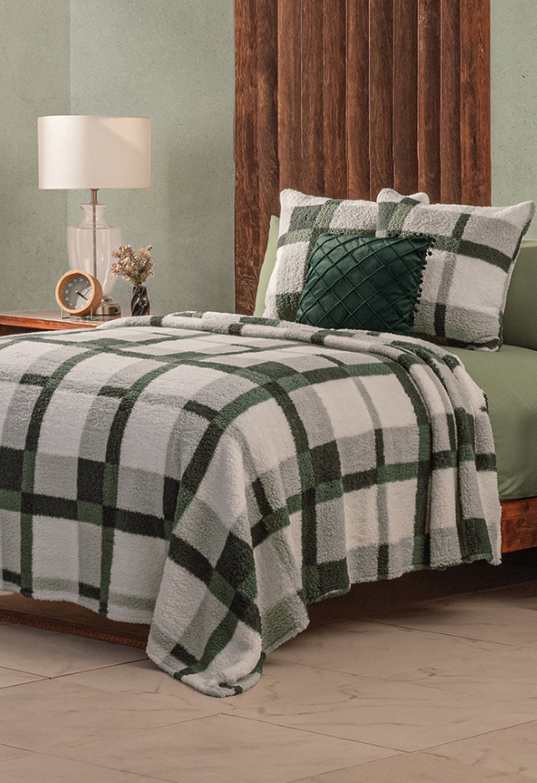 A neatly made bed features the Intima Hogar Juego de Cobertor Flannel Ligero Ottawa in green and white plaid, with matching pillows, beside a wood-paneled headboard. A bedside table with a lamp, clock, and flowers enhances the calm, neutral room.