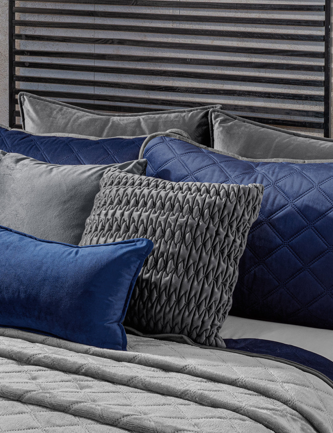 A neatly made bed showcases the Intima Hogar Juego de Cobertor Velvet Lovett Azul, with textured pillows—including a standout gray woven pillow and a blue velvet pillow—set against a dark slatted headboard.