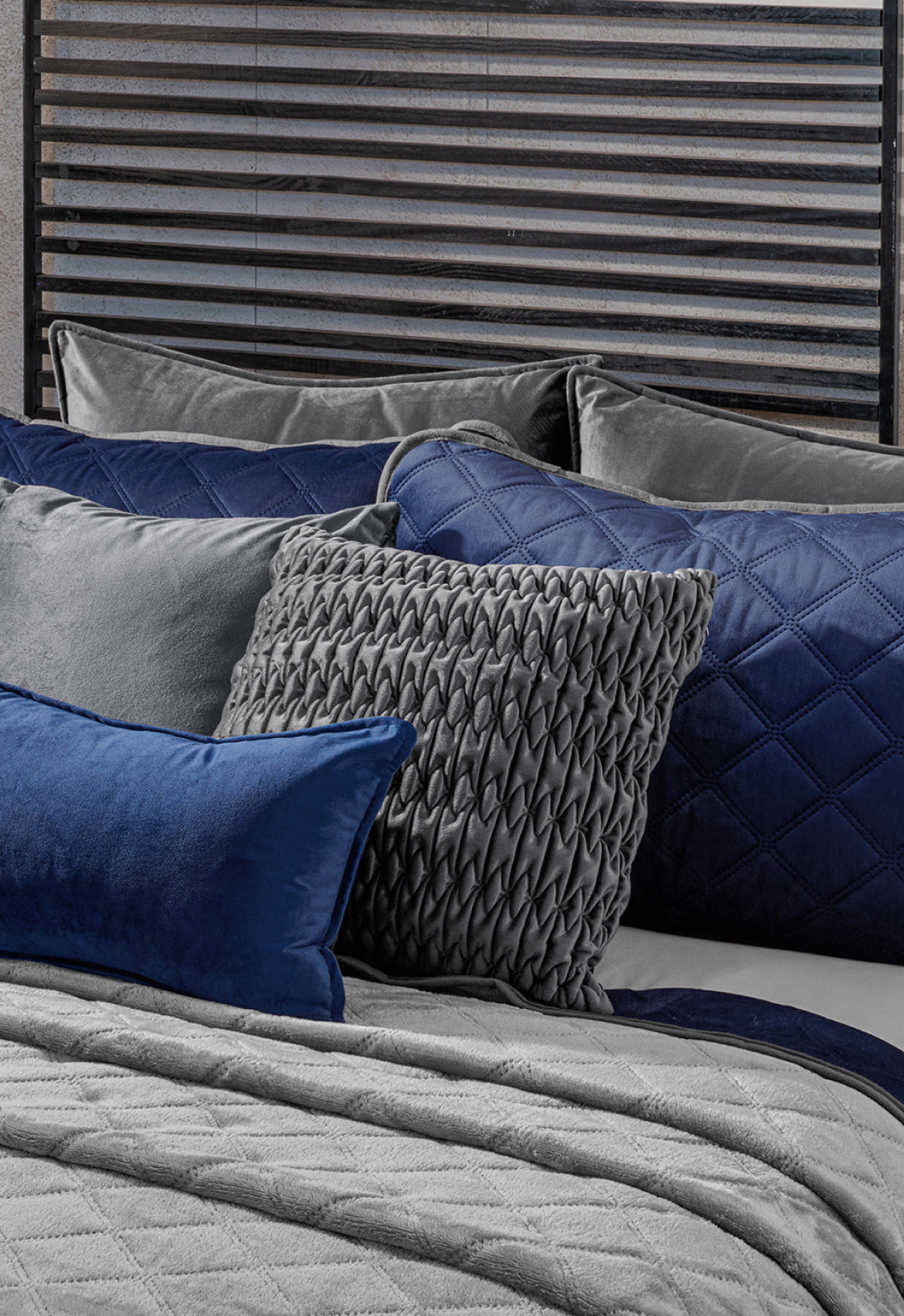 A neatly made bed showcases the Intima Hogar Juego de Cobertor Velvet Lovett Azul, with textured pillows—including a standout gray woven pillow and a blue velvet pillow—set against a dark slatted headboard.