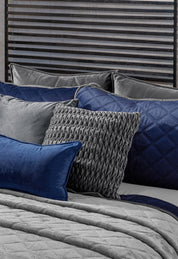 A neatly made bed showcases the Intima Hogar Juego de Cobertor Velvet Lovett Azul, with textured pillows—including a standout gray woven pillow and a blue velvet pillow—set against a dark slatted headboard.
