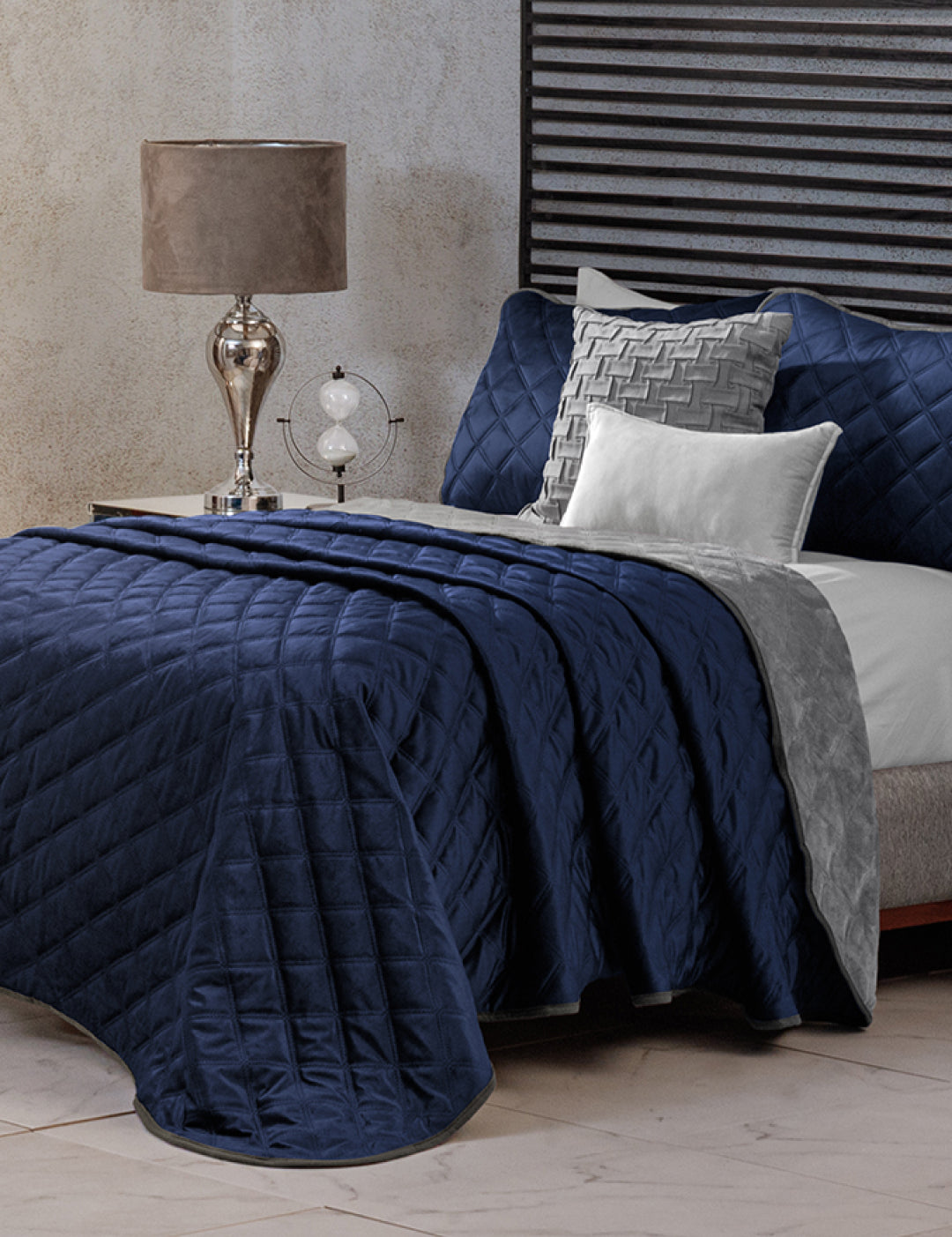 A neatly made bed features the Intima Hogar Juego de Cobertor Velvet Lovett Azul, complemented by gray and white accent pillows and a gray throw. A glass lamp and decorative globe rest on the bedside table, with a textured wall and striped headboard behind.
