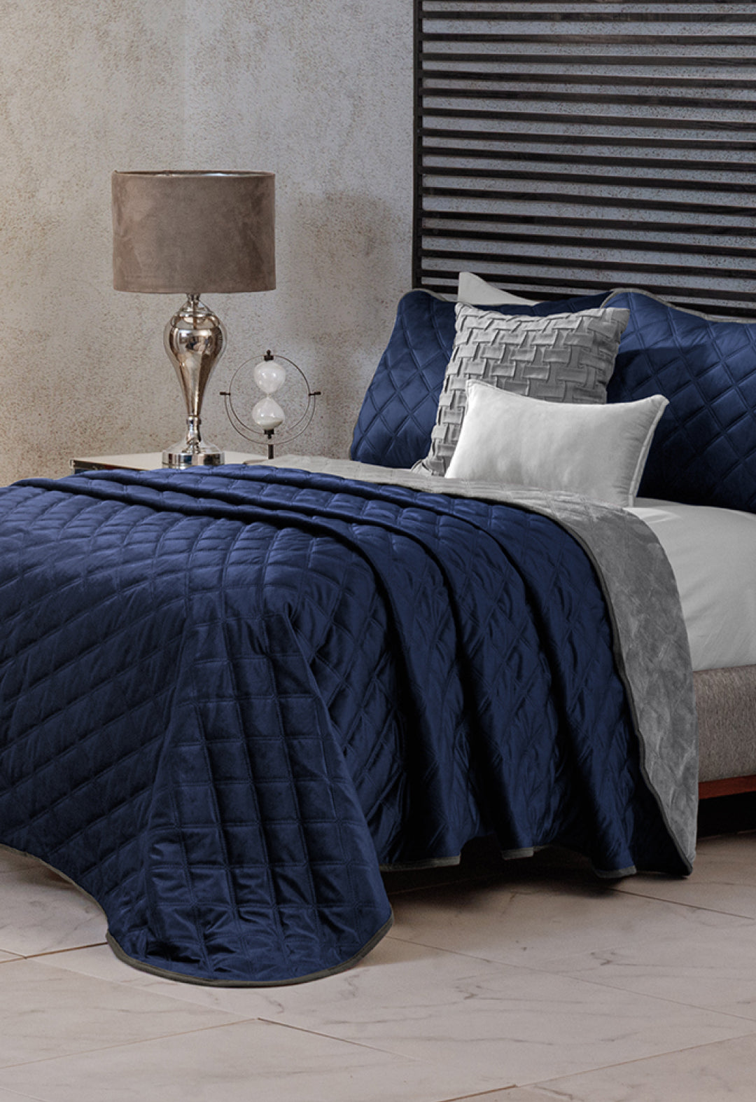 A neatly made bed features the Intima Hogar Juego de Cobertor Velvet Lovett Azul, complemented by gray and white accent pillows and a gray throw. A glass lamp and decorative globe rest on the bedside table, with a textured wall and striped headboard behind.