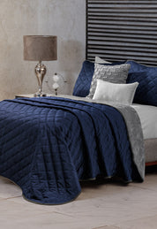 A neatly made bed features the Intima Hogar Juego de Cobertor Velvet Lovett Azul, complemented by gray and white accent pillows and a gray throw. A glass lamp and decorative globe rest on the bedside table, with a textured wall and striped headboard behind.