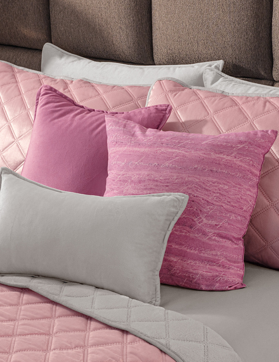 A neatly made bed showcases the Juego de Cobertor Velvet Lovett Rosa by Intima Hogar, including a pink quilted cobertor and plush pillows, all set against a cushioned brown headboard.