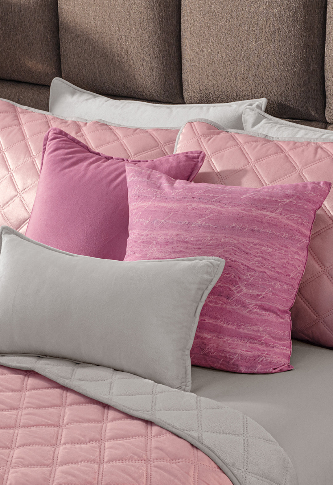 A neatly made bed showcases the Juego de Cobertor Velvet Lovett Rosa by Intima Hogar, including a pink quilted cobertor and plush pillows, all set against a cushioned brown headboard.