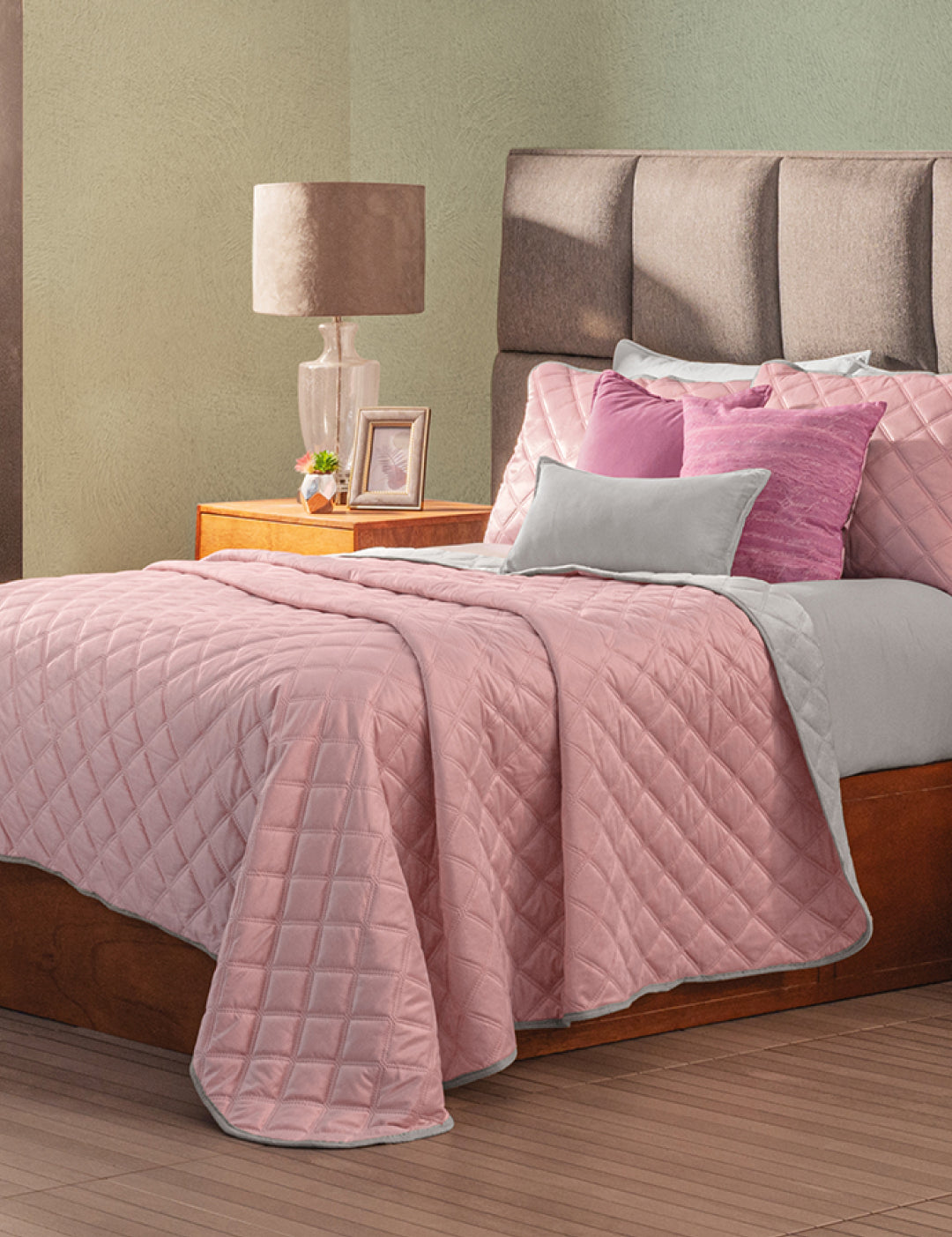 A neatly made bed features the Intima Hogar Juego de Cobertor Velvet Lovett Rosa with matching pillows. A wooden nightstand holding a lamp, photo frame, and decor stands beside the bed.