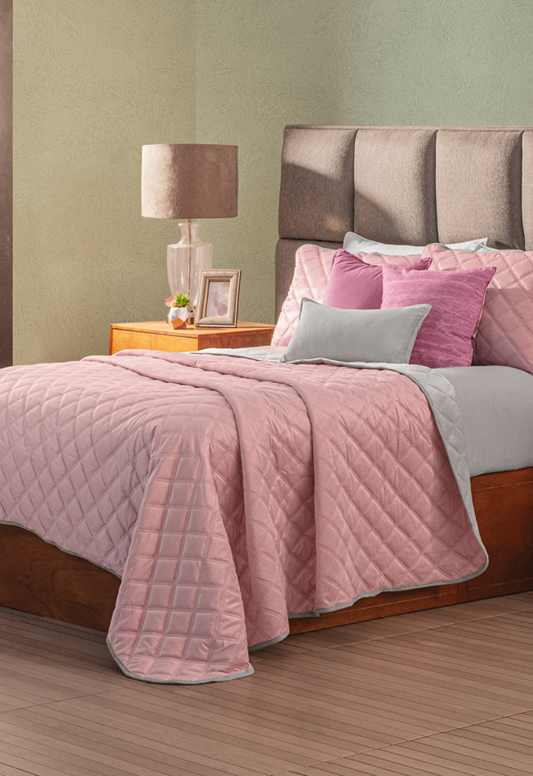 A neatly made bed features the Intima Hogar Juego de Cobertor Velvet Lovett Rosa with matching pillows. A wooden nightstand holding a lamp, photo frame, and decor stands beside the bed.