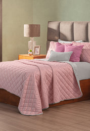 A neatly made bed features the Intima Hogar Juego de Cobertor Velvet Lovett Rosa with matching pillows. A wooden nightstand holding a lamp, photo frame, and decor stands beside the bed.