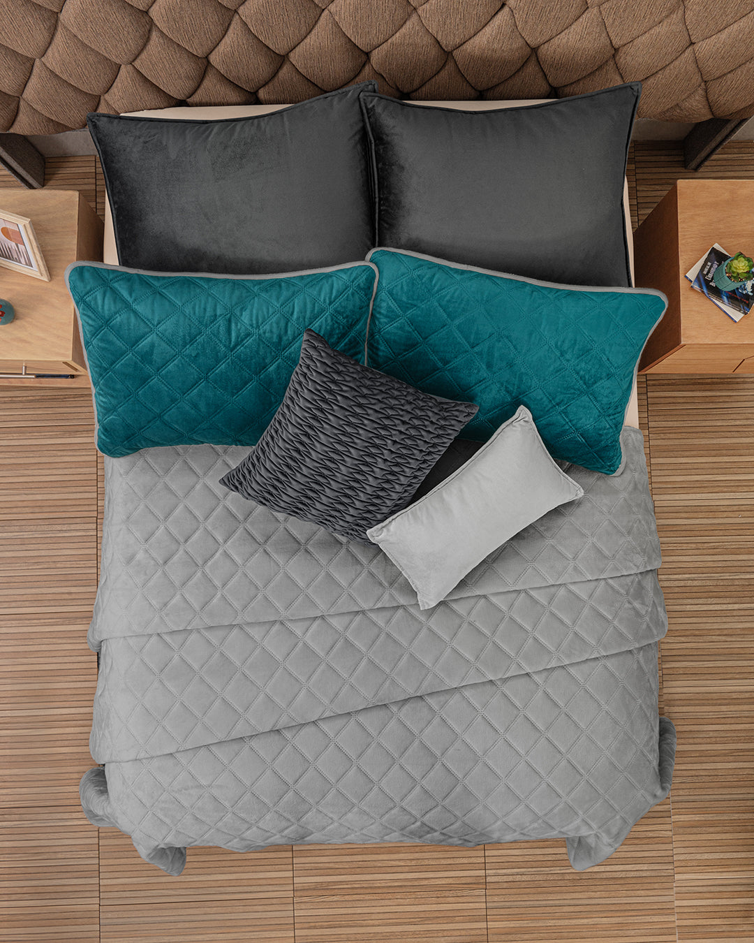 Overhead view of a bed with the Intima USA Funda de Almohada Ensueño Gris Oxford, a quilted gray comforter, black and teal pillows, and a textured gray cushion, set against a brown tufted headboard and wooden nightstands in the habitación.