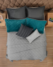 Overhead view of a bed with the Intima USA Funda de Almohada Ensueño Gris Oxford, a quilted gray comforter, black and teal pillows, and a textured gray cushion, set against a brown tufted headboard and wooden nightstands in the habitación.