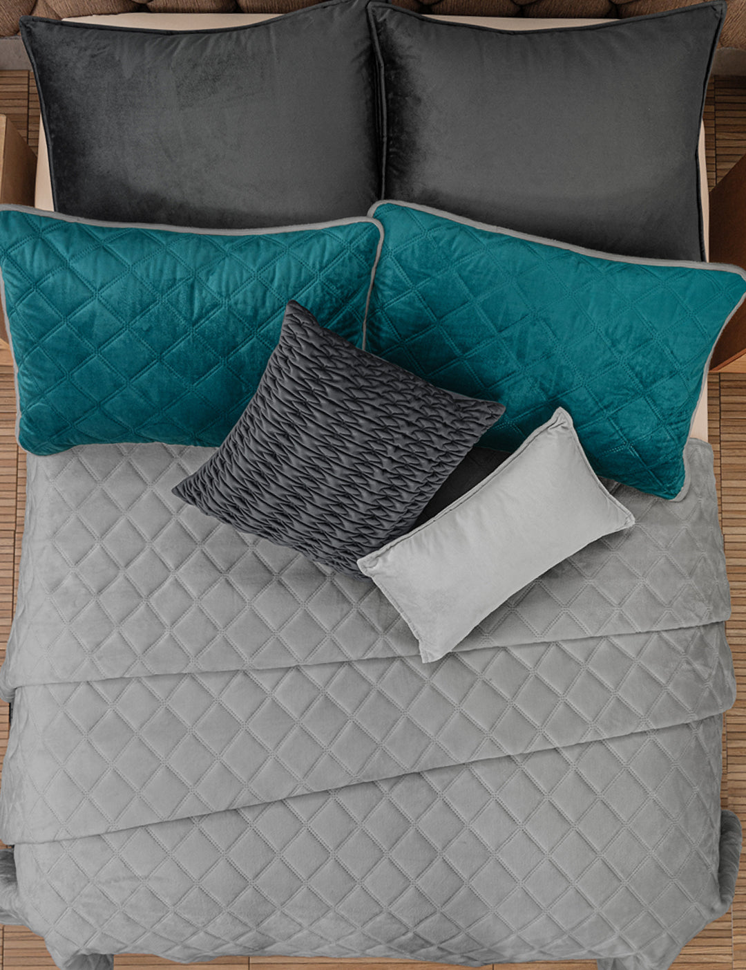 A neatly made bed with the Juego de Cobertor Velvet Lovett Verde by Intima Hogar, four large pillows in black and teal decorative covers, and two smaller pillows in gray and dark gray, all set on a wooden floor.