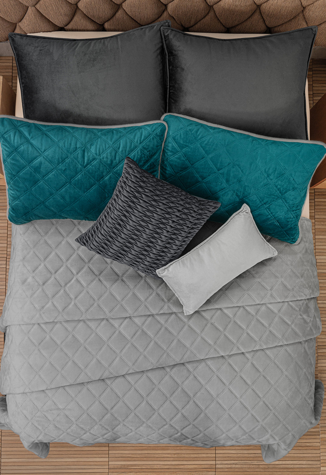 A neatly made bed with the Juego de Cobertor Velvet Lovett Verde by Intima Hogar, four large pillows in black and teal decorative covers, and two smaller pillows in gray and dark gray, all set on a wooden floor.