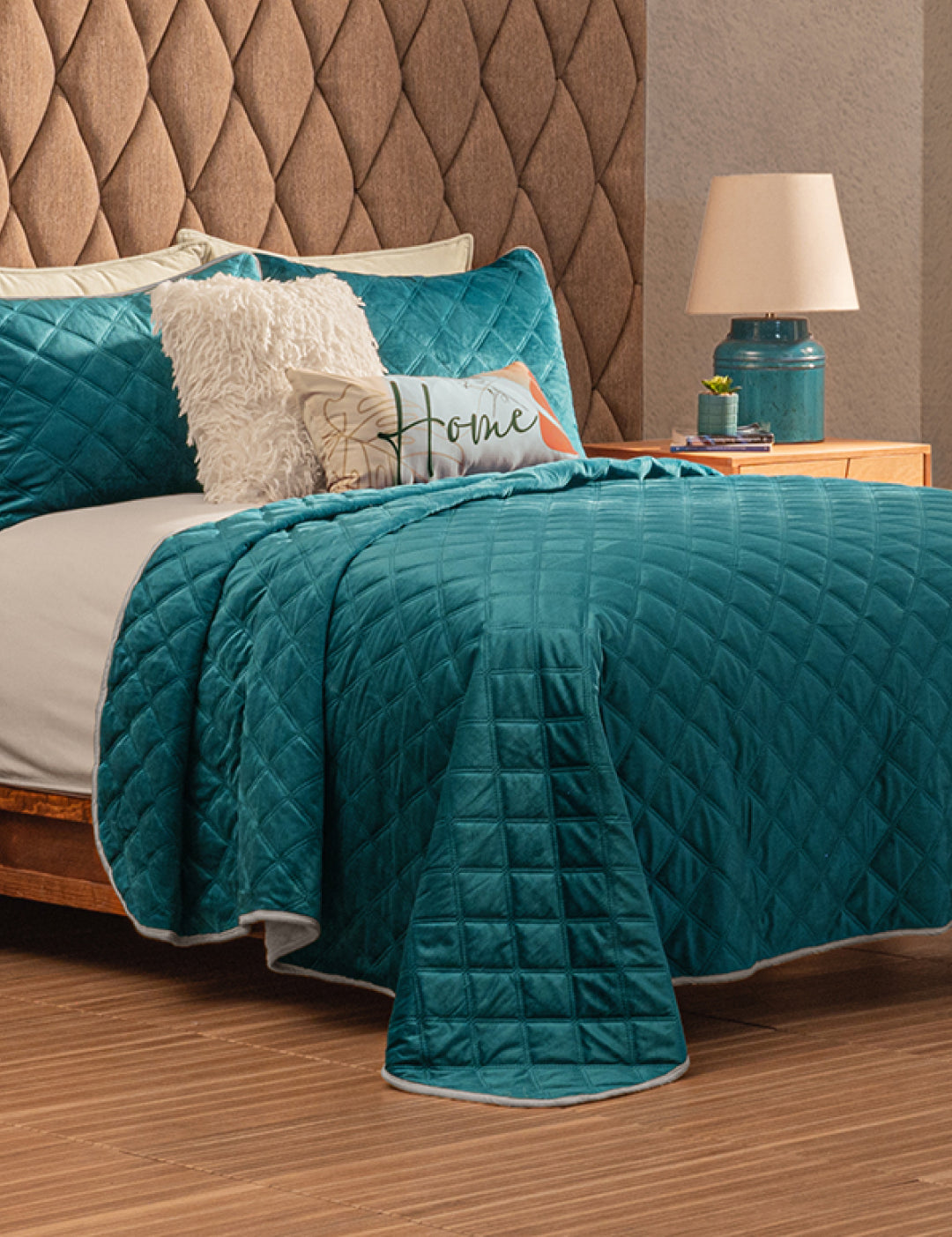 A neatly made bed features the Juego de Cobertor Velvet Lovett Verde by Intima Hogar, with white and teal pillows, a decorative Home pillow, and a fluffy cushion. The tufted headboard and teal lamp highlight the cobertor’s textura aterciopelada.