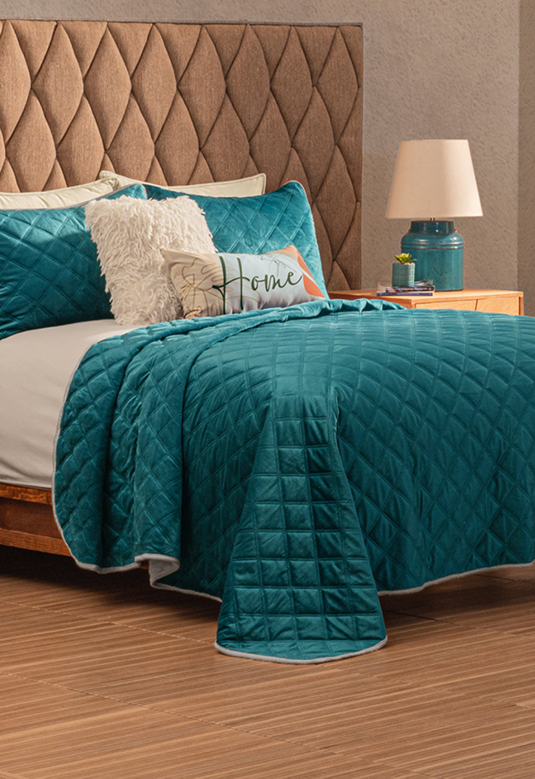 A neatly made bed features the Juego de Cobertor Velvet Lovett Verde by Intima Hogar, with white and teal pillows, a decorative Home pillow, and a fluffy cushion. The tufted headboard and teal lamp highlight the cobertor’s textura aterciopelada.