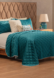 A neatly made bed features the Juego de Cobertor Velvet Lovett Verde by Intima Hogar, with white and teal pillows, a decorative Home pillow, and a fluffy cushion. The tufted headboard and teal lamp highlight the cobertor’s textura aterciopelada.
