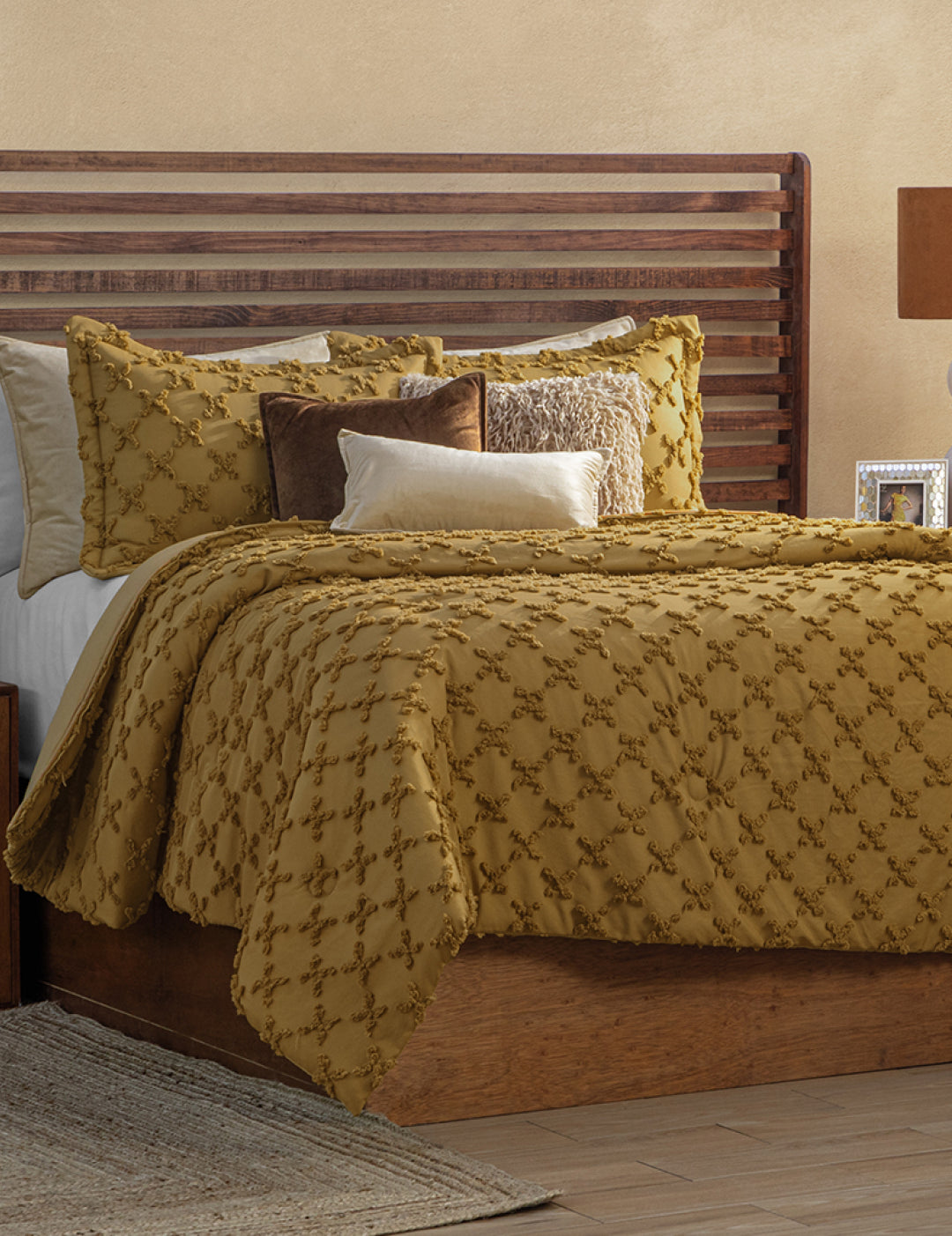 A neatly made bed with a wooden headboard features the Juego de Edredón Curry by Intima Hogar and decorative pillows in gold, white, and brown, all set on a wooden frame in a warm, neutral-toned room.