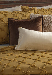 A close-up of a bed featuring the Juego de Edredón Curry by Intima Hogar, a mustard yellow chenille comforter with knotted patterns, paired with brown, shaggy cream, and soft ivory pillows.