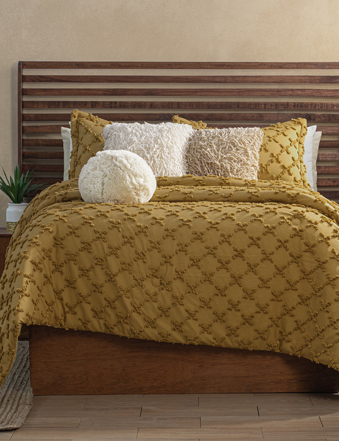 A neatly made bed features the Intima Hogar Juego de Edredón Curry, three decorative pillows (two square, one round) in white and beige, a wooden slatted headboard, and a small plant on the bedside table.