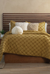 A neatly made bed features the Intima Hogar Juego de Edredón Curry, three decorative pillows (two square, one round) in white and beige, a wooden slatted headboard, and a small plant on the bedside table.