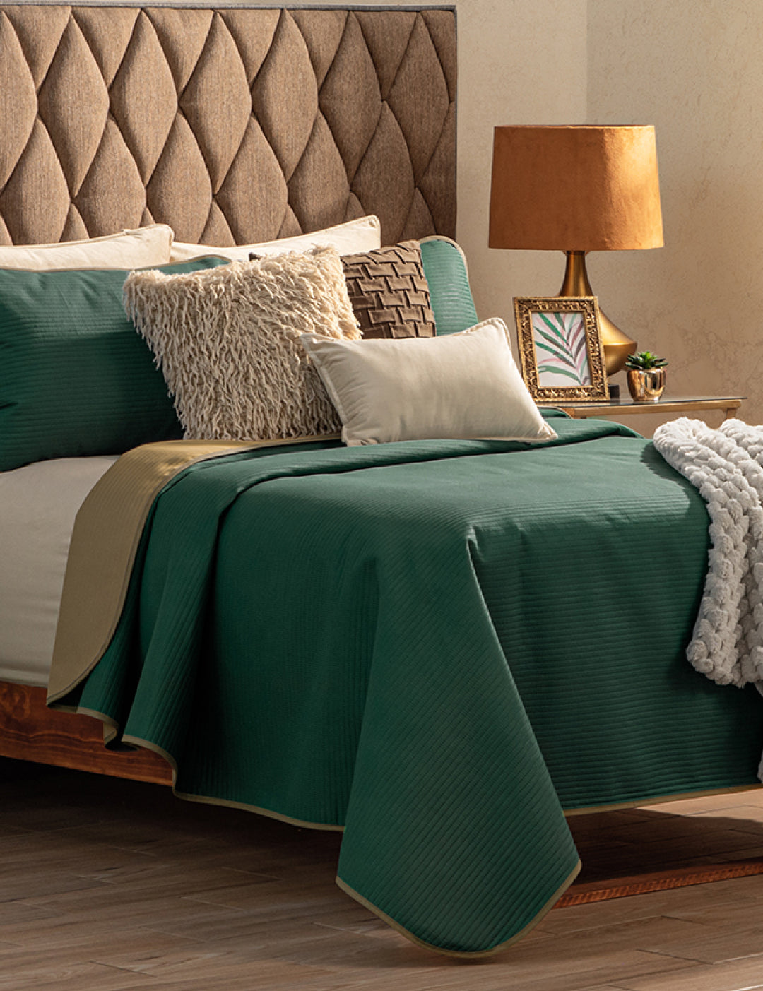 A neatly made bed with the Intima Hogar Juego de Edredón Ligero Honor, beige textured pillows, a beige knit throw, and a tan upholstered headboard. Next to it is a wooden nightstand with a lamp, framed photo, and small plant.