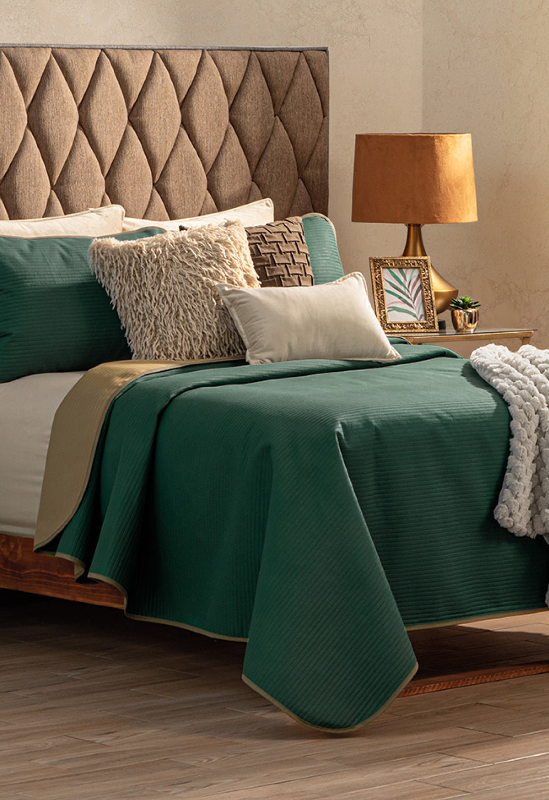 A neatly made bed with the Intima Hogar Juego de Edredón Ligero Honor, beige textured pillows, a beige knit throw, and a tan upholstered headboard. Next to it is a wooden nightstand with a lamp, framed photo, and small plant.