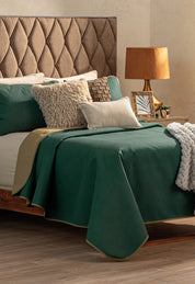 A neatly made bed with the Intima Hogar Juego de Edredón Ligero Honor, beige textured pillows, a beige knit throw, and a tan upholstered headboard. Next to it is a wooden nightstand with a lamp, framed photo, and small plant.