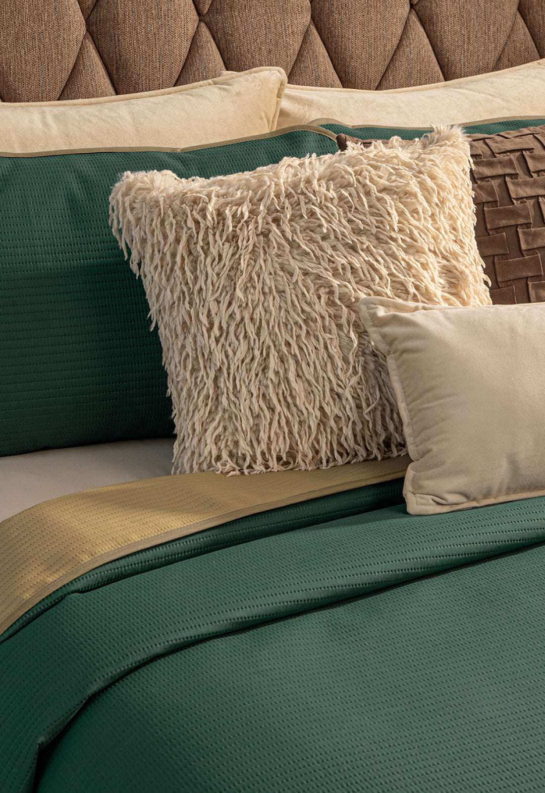 A neatly made bed features the Juego de Edredón Ligero Honor by Intima Hogar in green and gold, with decorative cream, brown woven, and light beige pillows. A tufted brown fabric headboard completes the look.