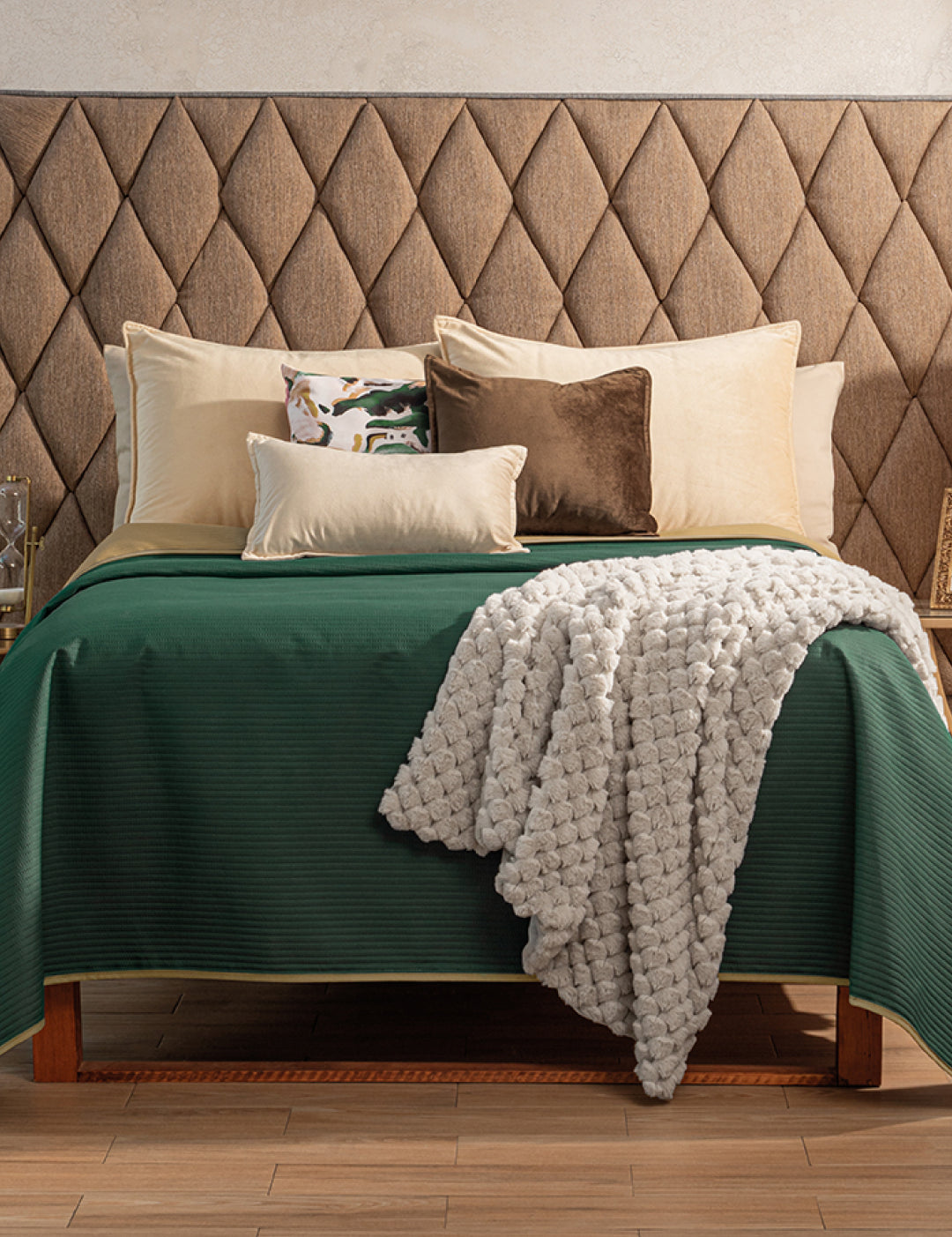 A neatly made bed showcases the Juego de Edredón Ligero Honor by Intima Hogar in green, accented with beige and brown pillows, a patterned cushion, and a textured white edredón capitonado. The diamond-patterned headboard and wooden floor complete the look.